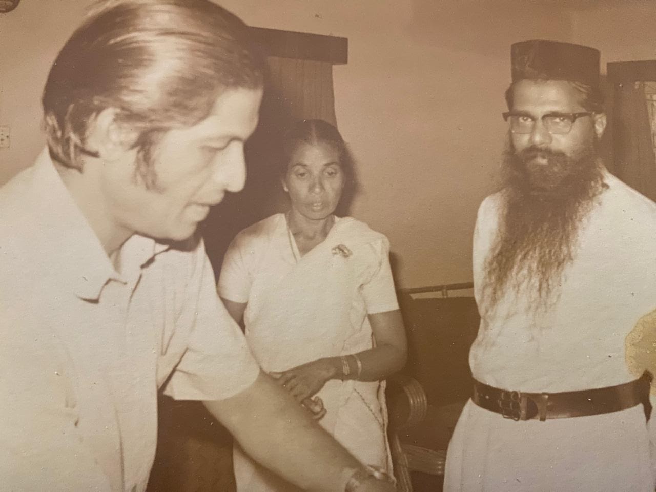 Indoor scene with a man leaning forward, a woman behind him, and another man with a long beard looking at the camera.