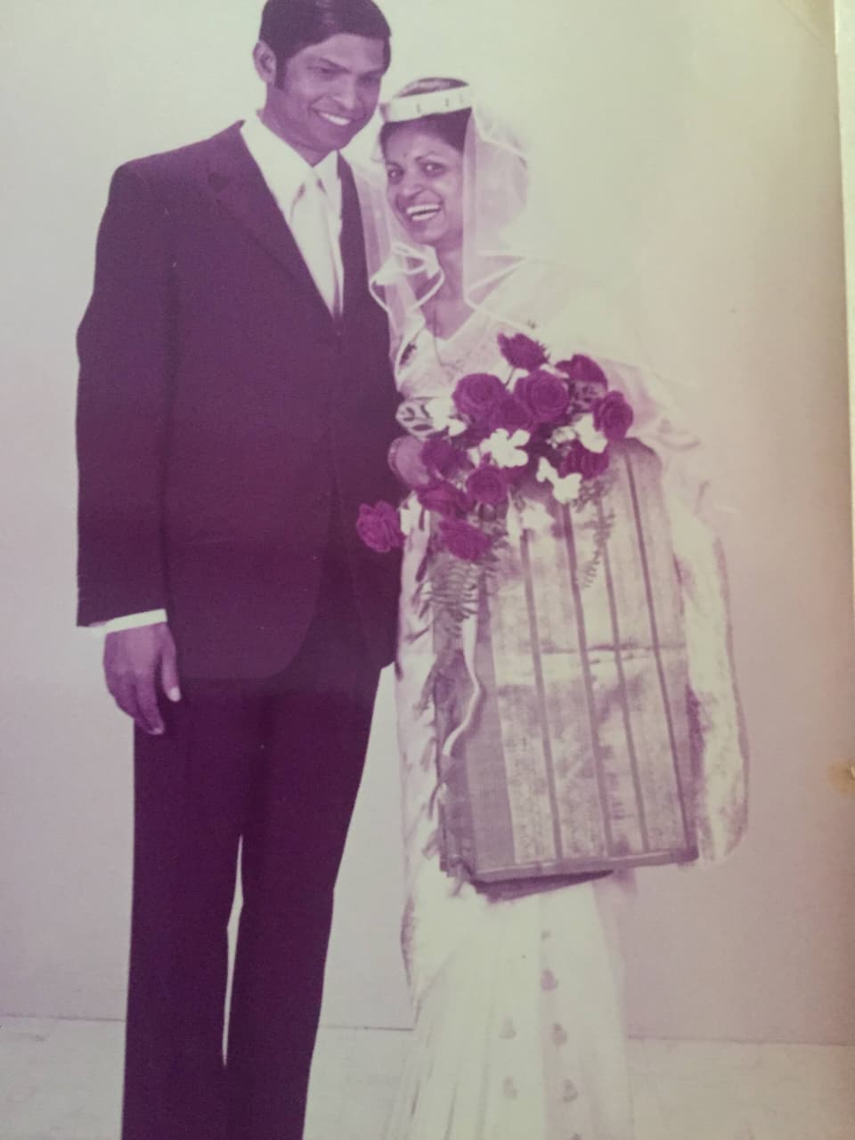 A man in a suit standing beside a woman in a wedding sari and veil, both smiling.